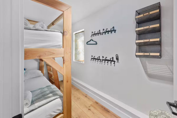 Small bright bedroom with natural wood bunk beds, white walls, palm-tree shaped wall hooks, a woven wall organizer, and light hardwood floors under a narrow window.