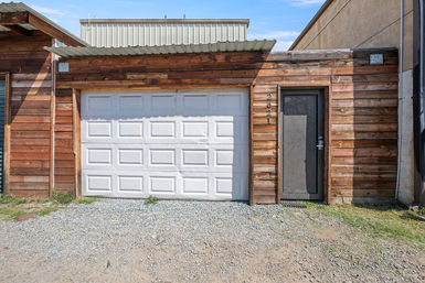 Rustic wood-clad building with a white paneled garage door, dark entry door, corrugated metal awning and gravel driveway on a sunny day.