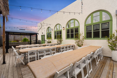 Cozy outdoor patio dining area at sunset with long banquet tables and white folding chairs on a wooden deck, string lights and green arched windows.