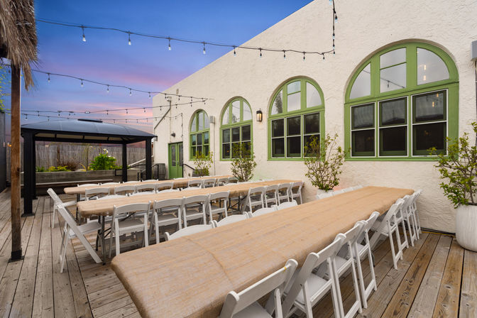Cozy outdoor patio dining area at sunset with long banquet tables and white folding chairs on a wooden deck, string lights and green arched windows.