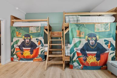 Wooden loft-style bunk beds with center ladder in a bright kids' bedroom, lower bunks hidden by colorful monkey-in-bathtub curtains, mint green accent wall and light wood floor.