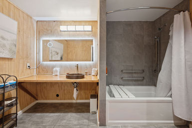 Interior accessible bathroom with wood vanity and round vessel sink, LED backlit mirror, gray tile floor and walls, and a bathtub-shower combo with grab bars and white curtain.