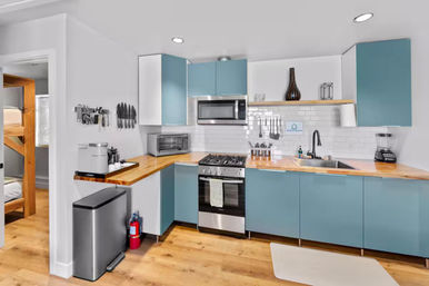 Bright modern kitchen with teal-blue cabinets and butcher-block countertops, stainless steel gas range and microwave, white subway tile backsplash, black faucet and sink, countertop appliances and knife rack, hardwood floors and bunk beds visible in the adjacent room.