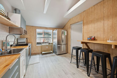 Bright modern kitchen with wood-paneled walls, stainless-steel refrigerator and appliances, wooden countertops and farmhouse sink, breakfast bar with four black metal stools, large window and light wood-look flooring.