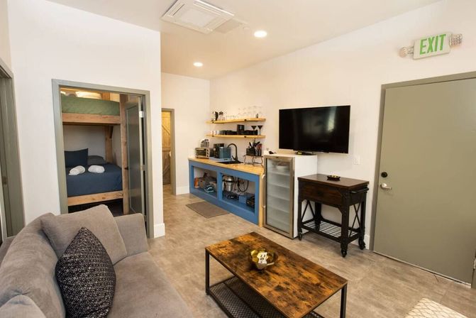 Cozy modern studio rental interior with gray sofa, wooden coffee table, wall-mounted TV, compact kitchenette with open shelves and mini fridge, and a bunk-bed nook.