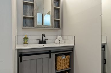 Small modern farmhouse bathroom vanity with gray wood cabinet and sliding barn-door, white countertop and subway-tile backsplash, black matte faucet, stacked white towels, woven basket and decorative soap dispenser