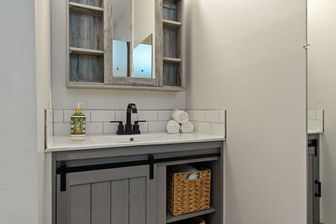 Small modern farmhouse bathroom vanity with gray wood cabinet and sliding barn-door, white countertop and subway-tile backsplash, black matte faucet, stacked white towels, woven basket and decorative soap dispenser