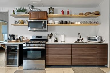 Modern kitchen with stainless steel gas range, over-the-range microwave, wine fridge and toaster oven; marble countertop, dark wood lower cabinets, black sink faucet, and natural wood floating shelves displaying dishes, glassware, bowls, plants and decorative items.