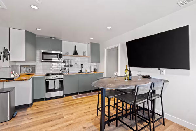 Modern open-plan apartment kitchen with gray cabinets, stainless steel stove and microwave, white subway tile backsplash and wood countertops, hardwood floors, round wooden dining table with bar stools, large wall-mounted flat-screen TV and wine glasses on the table.