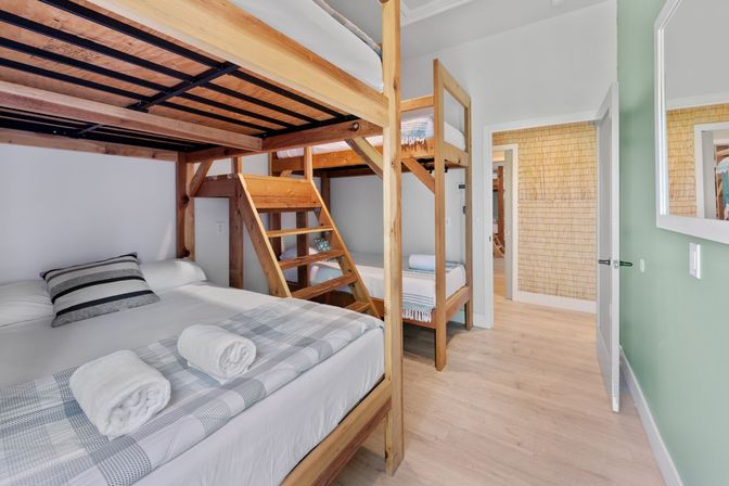 Coastal vacation rental bunk bedroom with natural wood bunk beds, rolled towels on the lower bed, light hardwood floors and a soft green accent wall.