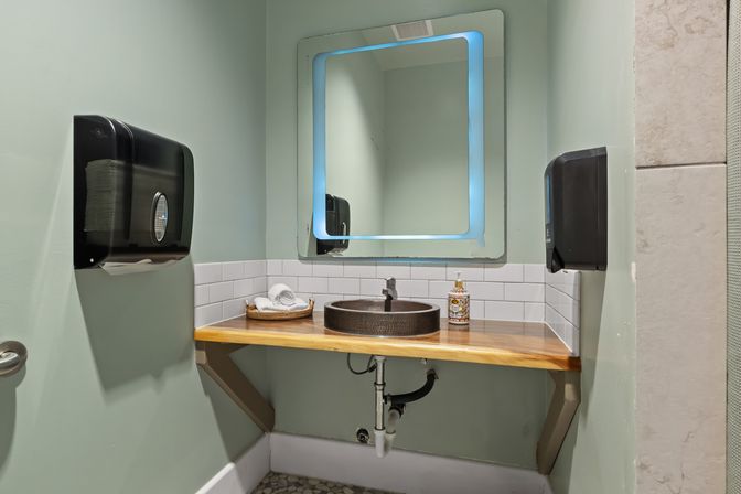 Modern restroom sink with a round metal vessel basin on a wood countertop, illuminated LED mirror, white subway-tile backsplash, wall-mounted paper towel dispensers, soap bottle and rolled towels.