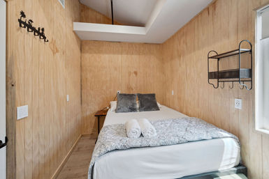 Cozy cabin-style wood-paneled bedroom with a double bed, gray throw pillows, patterned blanket, two rolled white towels, wall shelf and small side table.