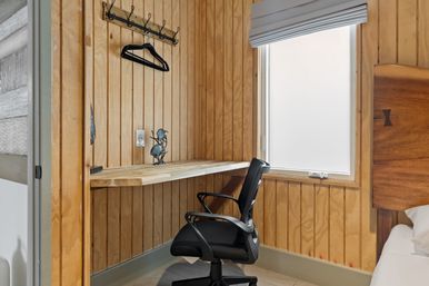 Cozy wood-paneled bedroom workspace with a floating wood desk, black swivel office chair, wall hooks with hangers, and a frosted window letting in soft natural light.
