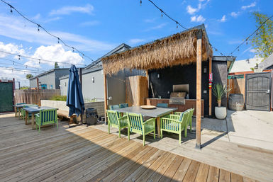 Sunny urban outdoor patio deck with a thatched tiki pergola, outdoor kitchen grill, long dining table surrounded by lime-green chairs and overhead string lights