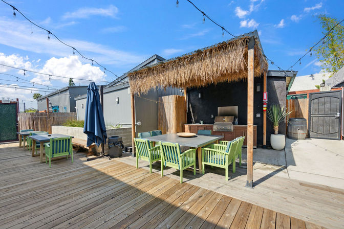 Sunny urban outdoor patio deck with a thatched tiki pergola, outdoor kitchen grill, long dining table surrounded by lime-green chairs and overhead string lights