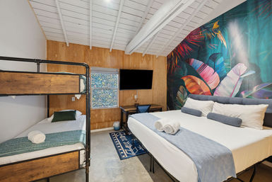 Cozy modern bedroom with vaulted white ceiling, vibrant tropical leaf mural accent wall, king bed with blue linens and bolsters, wooden bunk bed, flat-screen TV and small desk, rolled towels on the beds.