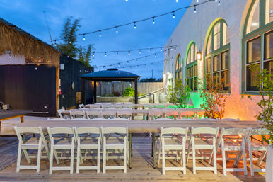 Dusk outdoor patio event space on a wooden deck with long rustic banquet tables and white folding chairs under string lights, arched stucco windows with colorful uplighting, a gazebo and thatched-roof bar area.