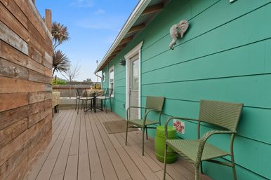 Sunny backyard deck beside a teal-sided cottage with two green wicker chairs, a small green side table, round bistro table near a wood privacy wall and potted palms under a blue sky.