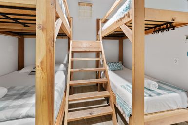Cozy shared bedroom with natural wood bunk beds and central ladder, crisp white linens, teal-striped throws and rolled towels in a bright compact space for families or groups.