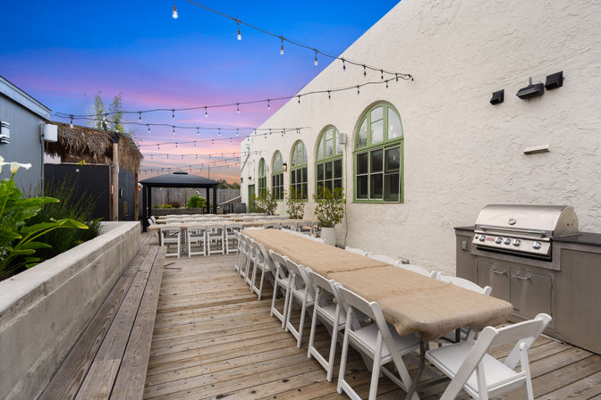 Outdoor wooden-deck patio with a long communal table and white folding chairs under string lights at sunset, arched green windows and a stainless grill.
