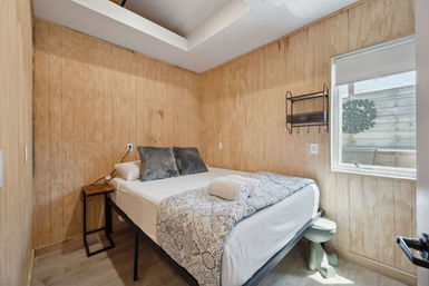 Cozy wood-paneled guest bedroom with elevated bed, patterned throw, rolled towels, gray accent pillows and a sunny window