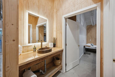 Cozy rustic-modern wood-paneled bathroom with illuminated square mirror, bronze vessel sink on a wooden vanity and an open door to a minimalist bedroom with concrete floors.