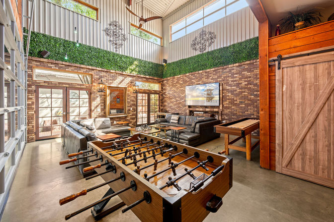 Bright industrial-loft game room with a foosball table in the foreground, shuffleboard and leather sectional by exposed brick walls, tall windows, high ceiling and a decorative green wall.