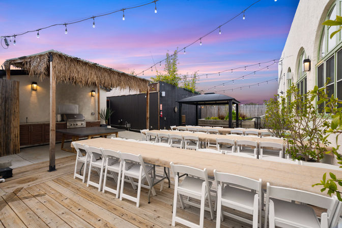 Cozy outdoor wooden deck dining area with long banquet tables and white folding chairs under string lights at sunset, thatched grill hut and small black gazebo