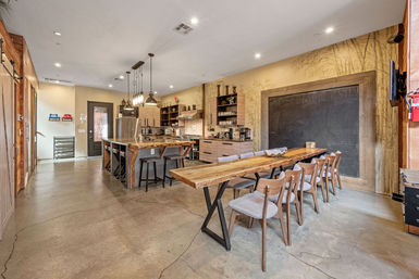 Open-concept industrial-chic communal kitchen and dining space with concrete floors, a long live-edge wooden table seating eight, a central island with bar stools, stainless steel appliances, pendant lighting, open shelving and a large framed chalkboard wall.