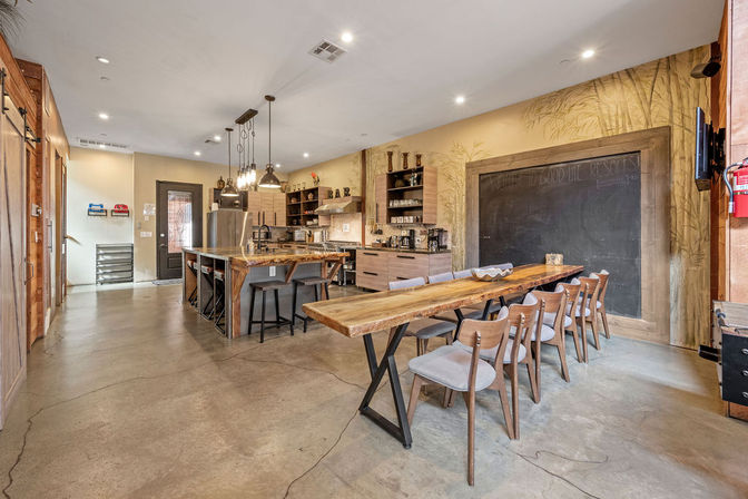 Open-concept industrial-chic communal kitchen and dining space with concrete floors, a long live-edge wooden table seating eight, a central island with bar stools, stainless steel appliances, pendant lighting, open shelving and a large framed chalkboard wall.