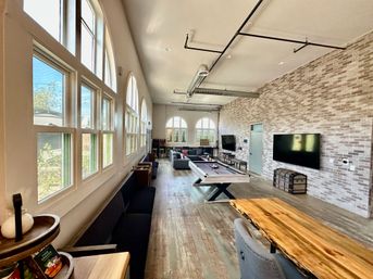 Bright, airy game room with tall arched windows, pool table, large wall-mounted TV, exposed ductwork, brick accent wall and wood plank floors.