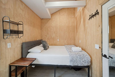 Cozy compact guest bedroom with light plywood walls, full bed with gray upholstered headboard, white sheets and patterned throw, rolled towel on the bed, metal wall shelf, small wooden side table and mirror on the door.