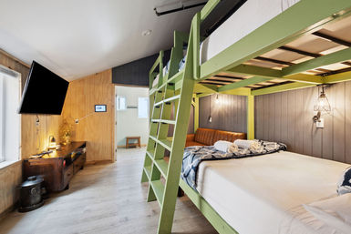 Cozy modern bedroom with a lime-green loft bunk, lower queen bed with rolled towels, leather bench, wood-paneled walls, wall-mounted TV and industrial pendant lights.