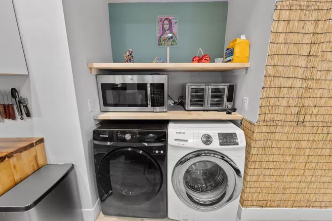 Compact apartment laundry nook with a black front-load dryer and white washer under wooden shelves holding a stainless microwave, toaster oven and yellow detergent jug, bamboo room divider and small trash can nearby.