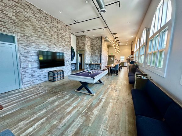 Sunlit community lounge with a pool table, long communal dining table, exposed brick wall with TV, arched windows, industrial ceiling and worn wooden floors.