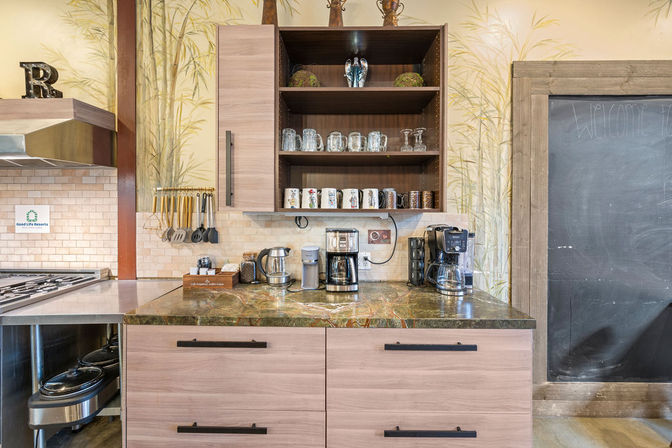 Cozy communal kitchen coffee station with two drip coffee makers, electric kettle, mugs and glassware on open wooden shelves, marble countertop, utensil rack, tiled backsplash and large chalkboard wall.