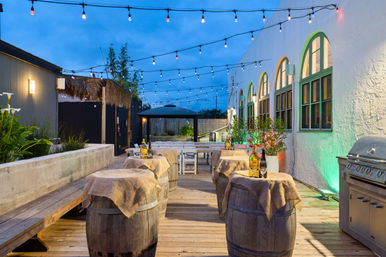 Evening outdoor patio on a wooden deck with hanging string lights, rustic wine-barrel tables draped in burlap topped with wine bottles, arched windows on a white building and a stainless grill — cozy alfresco dining setup.