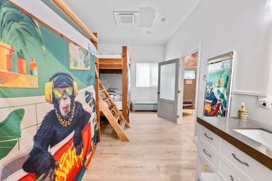 Bright modern bunk room with wooden loft bunks and ladder, playful colorful curtain showing a chimpanzee in headphones and sunglasses, white vanity with sink and mirror, light wood floors, and an open door to an adjacent room with a window.