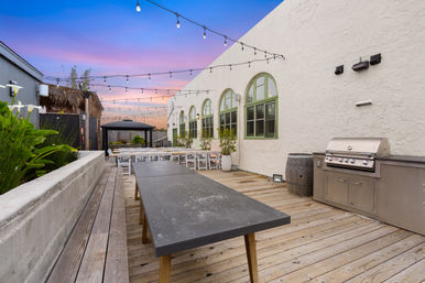 Rooftop wooden deck with long communal tables and benches, hanging string lights, stainless steel grill, arched green-trim windows, potted plants and a gazebo under a pink-purple sunset.