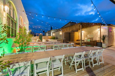 Outdoor evening dining patio at twilight with long wooden communal tables, white folding chairs, twinkling string lights, potted plants, and a thatched grill station beside a stucco building.
