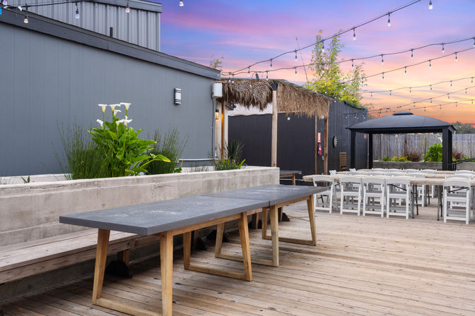 Rooftop patio with wooden deck, long concrete-topped communal table, white folding chairs, thatched pergola and string lights against a pink-purple sunset