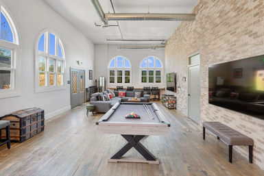 Sunlit urban loft game room with a central pool table on hardwood floors, cozy sectional sofa, arched windows, exposed ductwork and a brick accent wall with wall-mounted TV.