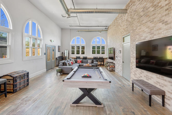 Sunlit urban loft game room with a central pool table on hardwood floors, cozy sectional sofa, arched windows, exposed ductwork and a brick accent wall with wall-mounted TV.