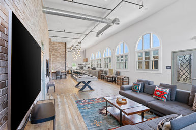 Bright industrial loft living room and open kitchen with high ceilings, exposed ductwork, arched windows, gray sectional with colorful pillows, wood coffee table on a patterned rug, and a central pool table on reclaimed wood floors.
