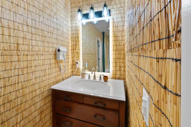 Small bathroom vanity with white sink and wooden drawers, illuminated mirror with three-bulb light, bamboo reed walls and wall-mounted hair dryer — cozy tropical-style washroom.