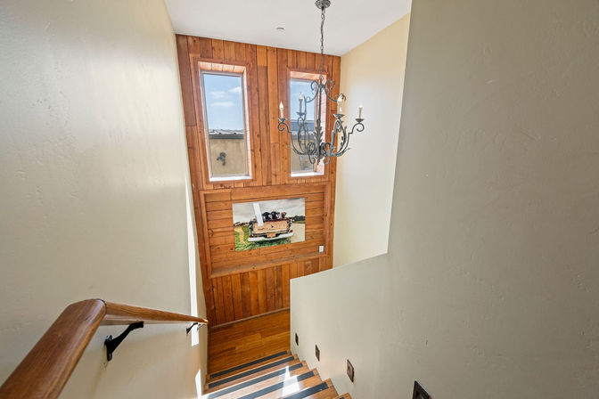 Sunlit interior staircase with oak handrail, wooden accent wall featuring two tall windows and a framed boat painting, ornate iron chandelier and hardwood steps.