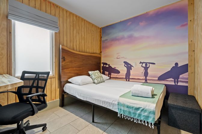 Cozy beach-themed guest room with wood-paneled walls, single bed with green throw and towel, surf-silhouette sunset mural, desk and black office chair by window