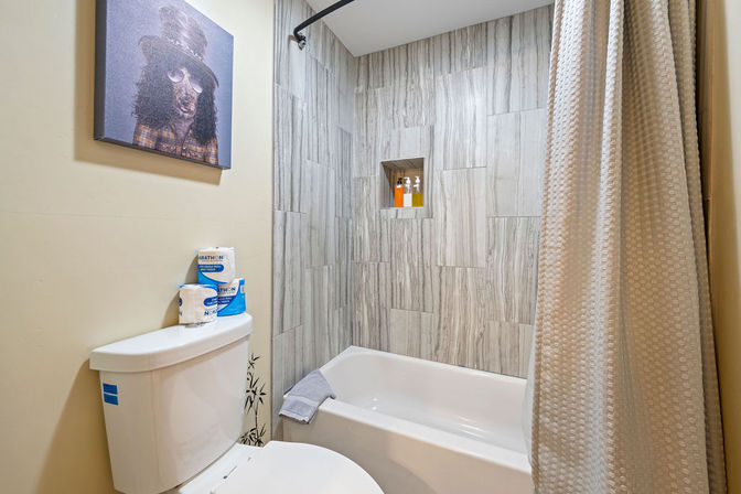 Small modern bathroom with bathtub-shower, vertical gray stone-look tile and recessed niche with shampoo bottles, white toilet topped with spare toilet paper, beige textured shower curtain and quirky dog portrait on the wall.