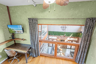 Mezzanine loft home office with wooden desk, swivel chair and wall-mounted TV, green textured walls and curtains, overlooking a sunlit open atrium with exposed brick and a faux-hedge accent.