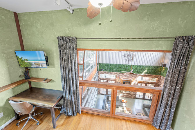 Mezzanine loft home office with wooden desk, swivel chair and wall-mounted TV, green textured walls and curtains, overlooking a sunlit open atrium with exposed brick and a faux-hedge accent.